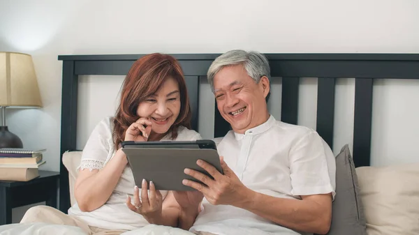 Asian senior couple using tablet at home. Asian Senior Chinese grandparents, husband and wife happy after wake up, watching movie lying on bed in bedroom at home in the morning concept.