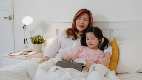 Asian grandmother using tablet read fairy tales to granddaughter at home. Senior Chinese, grandma happy relax with young girl before bedtime lying on bed in bedroom at home at night concept.