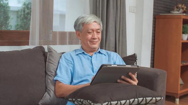 Asian senior men using tablet at home. Asian Senior Chinese male search information about how to good health on internet while lying on sofa in living room at home concept.