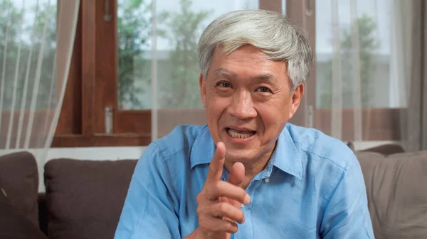 Asian senior man video call at home. Asian senior older Chinese male using mobile phone video call talking with family grandchild kids while lying on sofa in living room at home concept.