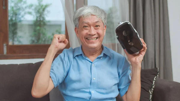 Asian senior men play games at home. Asian senior older Chinese male happy fun and virtual reality, VR playing games while lying sofa in living room at home concept.