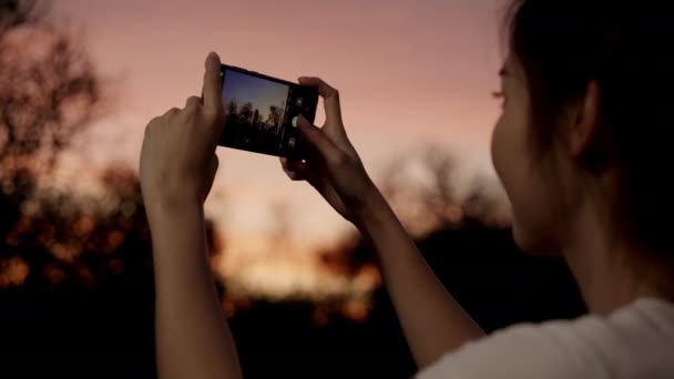 年轻快乐的亚洲女孩拿着手机 欢快地放松下来 在公园的森林树上拍一张和平落日的照片 美丽的自然景观日落 带着智能手机概念的旅行度假 — 图库视频影像