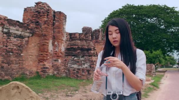若いアジアのバックパッカーの女性のブロガーカメラと観光客は 古い町でプラスチックボトルの中に休息と飲料水を取る塔の前に疲れてスタンドを感じる ライフスタイル観光旅行休日のコンセプト — ストック動画