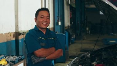 Profesyonel araba tamircisi kameraya bakıyor ve tamirhanede gülümsüyor. Geceleri tamirci garajında araba tamir eden Asyalı yetenekli bir adam. Araba servisi bakımı.
