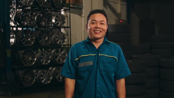 カメラを見て 修理サービスステーションで笑みを浮かべてプロの車の整備士 夜のメカニズムガレージで均一な固定車の熟練したアジアの男 車のメンテナンス — ストック動画