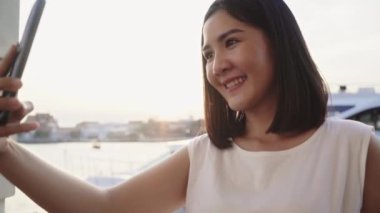 Mutlu Asyalı genç kadın blogger turist günlük stilde kullan akıllı telefon kullan selfie güzel günbatımı nehir manzaralı liman şehir kasabası, Lifestyle turizm tatil konsepti.