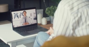 Bilgisayarlı dizüstü bilgisayar kullanan genç Asyalı kız video konferans görüşmesinde evdeki oturma odasında uzman doktorla hastalıktan bahsediyor. Coronavirus konsepti için sosyal uzaklık, karantina.