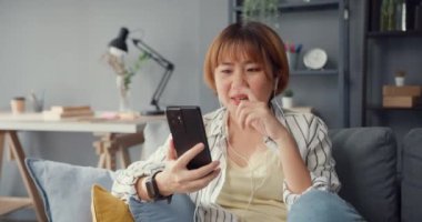 Genç Asyalı bayan, evdeki oturma odasındaki kanepede ailesiyle akıllı telefon görüşmeleri yapıyor. Covid-19 tecrit, sosyal uzaklık, koronavirüs önleme karantinası sırasında uzaktan evden çalışma.