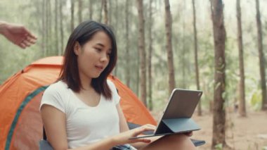 Genç Asyalı kampçılar ormanda çadırın yanında tablet kullanıyorlar. Erkek ve kadın gezgin bir yaz günü kamp alanında dinlenip sohbet ediyorlar. Açık hava aktivitesi, macera seyahati veya tatil tatili.