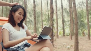Genç Asyalı kampçılar ormanda çadırın yanında tablet kullanıyorlar. Erkek ve kadın gezgin bir yaz günü kamp alanında dinlenip sohbet ediyorlar. Açık hava aktivitesi, macera seyahati veya tatil tatili.