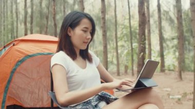 Genç Asyalı kampçılar ormanda çadırın yanında tablet kullanıyorlar. Erkek ve kadın gezgin bir yaz günü kamp alanında dinlenip sohbet ediyorlar. Açık hava aktivitesi, macera seyahati veya tatil tatili.