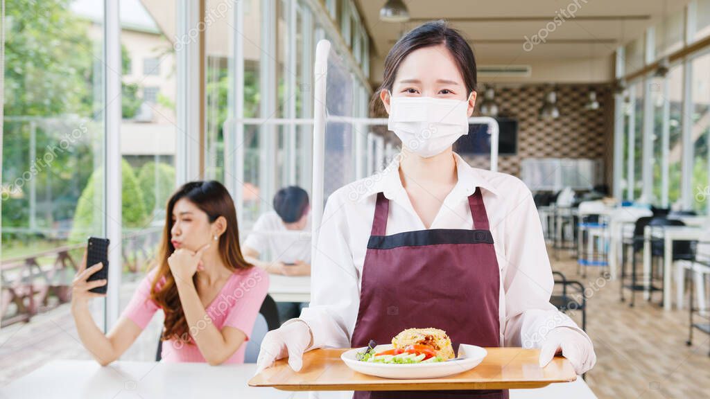 Young Asia female restaurant staff wearing protective face mask holding food tray to serving meal to customer with customer in background. Lifestyle new normal restaurant after corona virus concept.