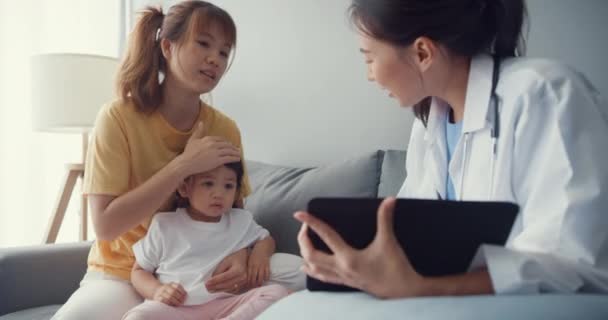 年轻的亚洲女儿科医生和使用数码平板电脑与快乐的妈妈分享健康测试的好消息的小女孩坐在沙发上 医疗保险 回家看病人 — 图库视频影像
