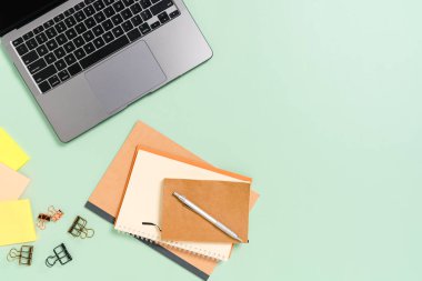 Çalışma masasının yaratıcı düz yatay fotoğrafı. Üst kattaki ofis masasında dizüstü bilgisayarı, kahve fincanı ve pastel yeşil arka planda açık siyah bir defter var. Üst görünüm, fotokopi alanı fotoğrafçılığıyla aynı..