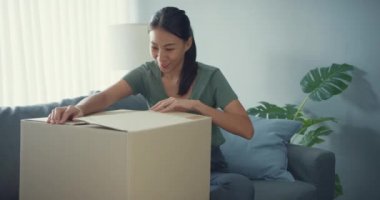 Mutlu Asyalı bayan karton kutu paketi açıyor. Heyecan verici ve evdeki oturma odasındaki çevrimiçi kıyafet ürünlerinin kalitesiyle uyumlu olmaya çalışıyor. Çevrimiçi alışveriş ve dağıtım kavramı.