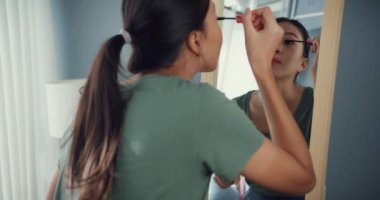 Gündelik kıyafetli güzel Asyalı kadın, sabahleyin dışarı çıkmadan önce yatak odasındaki aynanın önünde kirpiğine rimel sürmüş. Gülümseyen genç kadın makyaj yapıyor ve aynaya bakıyor..