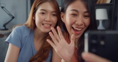 Mutlu gülümseyen Asyalı genç kadınlar rahatlamak için evdeki oturma odasında akıllı telefon görüntülerini kullanıyorlar. Neşeli Ev Arkadaşı Kadınlar, ev konseptinde yaşam tarzı bir kadınla arkadaş ve aile ile video konferansı..