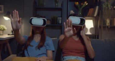 Çekici Asyalı kadınlar, karanlık bir gecede evdeki kanepe oturma odasında sanal gerçeklik gözlüğü kulaklığı sitesiyle online alışveriş yapmaktan zevk alırlar. Film saati için VR gözlüklü kulaklık kullanılıyor.