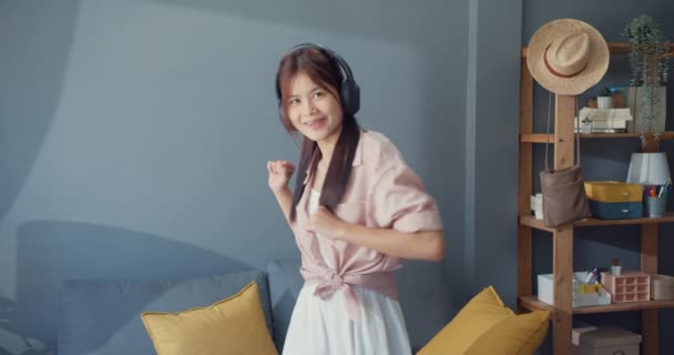 快乐无忧无虑的年轻亚洲女孩戴着耳机听音乐 独自在客厅里跳舞 孤立的社会距离头孢病毒流行的概念 享受自由和积极的生活方式概念 — 图库视频影像