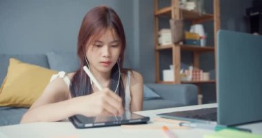 Young Asia girl teenager with casual shirt wear earphone use laptop learn online write lecture in laptop in living room at house. Isolate education online e-learning coronavirus pandemic concept.