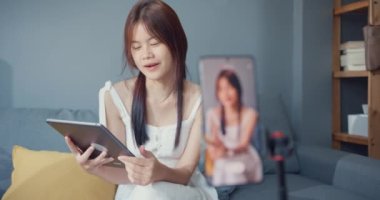Happy young Asian girl blogger in front of phone camera use tablet enjoy question answer with follower in living room at home. Blogger activity lifestyle, Social distance coronavirus pandemic concept.