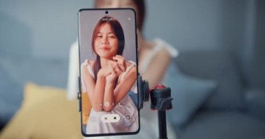 Happy young Asian girl blogger in front of phone camera use tablet enjoy question answer with follower in living room at home. Blogger activity lifestyle, Social distance coronavirus pandemic concept.