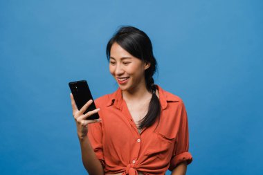 Genç Asyalı bayan telefonu olumlu bir ifadeyle kullanıyor, geniş bir şekilde gülümsüyor, gündelik giysiler içinde mutluluğu hissediyor ve mavi arka planda tek başına duruyor. Mutlu, mutlu, mutlu kadın başarıya sevinir..