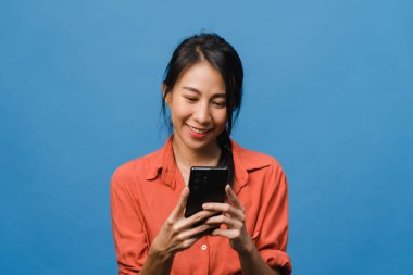 Genç Asyalı bayan telefonu olumlu bir ifadeyle kullanıyor, geniş bir şekilde gülümsüyor, gündelik giysiler içinde mutluluğu hissediyor ve mavi arka planda tek başına duruyor. Mutlu, mutlu, mutlu kadın başarıya sevinir..