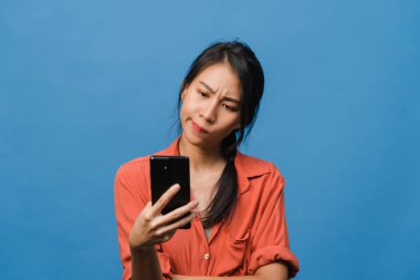 Genç Asyalı bayan telefonu olumlu bir ifadeyle kullanıyor, geniş bir şekilde gülümsüyor, gündelik giysiler içinde mutluluğu hissediyor ve mavi arka planda tek başına duruyor. Mutlu, mutlu, mutlu kadın başarıya sevinir..