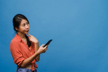 Genç Asyalı bayan telefonu olumlu bir ifadeyle kullanıyor, geniş bir şekilde gülümsüyor, gündelik giysiler içinde mutluluğu hissediyor ve mavi arka planda tek başına duruyor. Mutlu, mutlu, mutlu kadın başarıya sevinir..