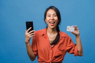 Genç Asyalı bayan telefon ve kredi kartını olumlu ifadeli kullanıyor, geniş bir gülümseme sergiliyor, günlük kıyafetler giyiyor ve mavi arka planda tek başına duruyor. Mutlu, mutlu, mutlu kadın başarıya sevinir..