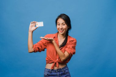 Genç Asyalı bayan kredi kartını pozitif bir ifadeyle gösteriyor, geniş bir şekilde gülümsüyor, günlük giysiler içinde mutluluğu hissediyor ve mavi arka planda tek başına duruyor. Yüz ifadesi kavramı.