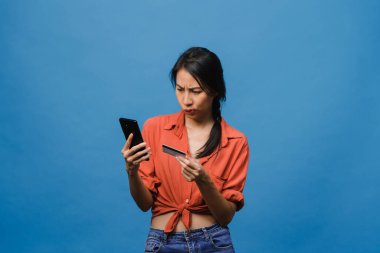 Genç Asyalı bayan telefon ve kredi kartı kullanıyor, olumsuz ifadeli, heyecanlı çığlıklar atıyor, duygusal kızgınlıkla ağlıyor ve mavi arka planda tek başına duruyor. Yüz ifadesi kavramı.
