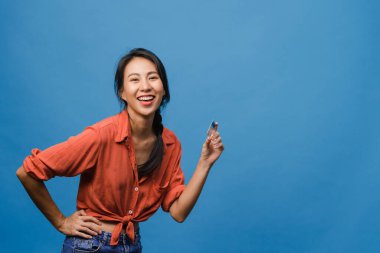 Genç Asyalı bayan kredi kartını pozitif bir ifadeyle gösteriyor, geniş bir şekilde gülümsüyor, günlük giysiler içinde mutluluğu hissediyor ve mavi arka planda tek başına duruyor. Yüz ifadesi kavramı.