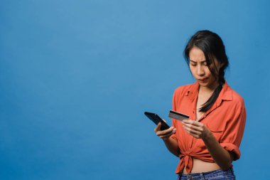 Genç Asyalı bayan telefon ve kredi kartı kullanıyor, olumsuz ifadeli, heyecanlı çığlıklar atıyor, duygusal kızgınlıkla ağlıyor ve mavi arka planda tek başına duruyor. Yüz ifadesi kavramı.