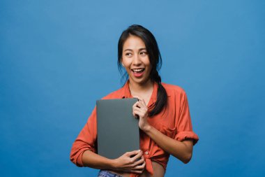 Genç Asyalı bayan dizüstü bilgisayarı olumlu bir ifadeyle tutuyor, geniş bir şekilde gülümsüyor, günlük giysiler içinde mutluluğu hissediyor ve mavi arka planda tek başına duruyor. Mutlu, mutlu, mutlu kadın başarıya sevinir..