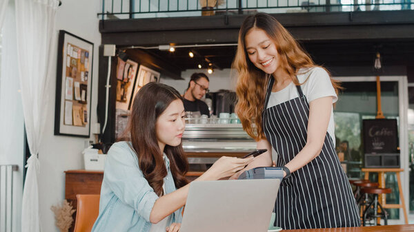 Young Asian freelance women pay contactless at coffee shop. Asian happy girl barista waiter wear gray apron holding credit card reader machine for customer using mobile phone scan pay in cafe.