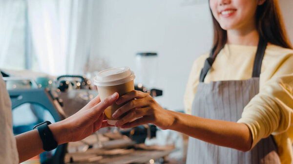 Young Asia female barista serving take away hot coffee paper cup to consumer standing behind bar counter at cafe restaurant. Owner small business, food and drink, service mind concept.