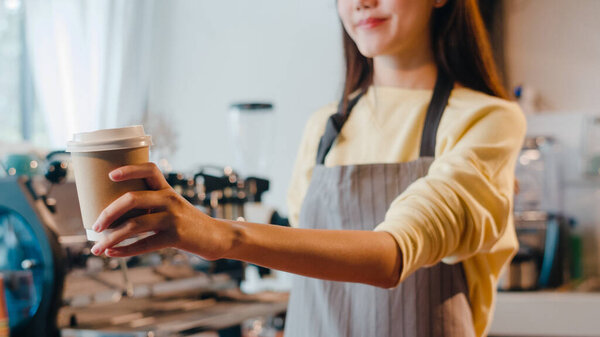 Young Asia female barista serving take away hot coffee paper cup to consumer standing behind bar counter at cafe restaurant. Owner small business, food and drink, service mind concept.