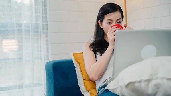 若いビジネスフリーランスのアジア人女性は 自宅のリビングルームでリラックスしたときにソファに横たわっている間 ソーシャルメディアとコーヒーを飲みながらラップトップで作業しています 家のコンセプトでライフスタイルの女性 — ストック写真