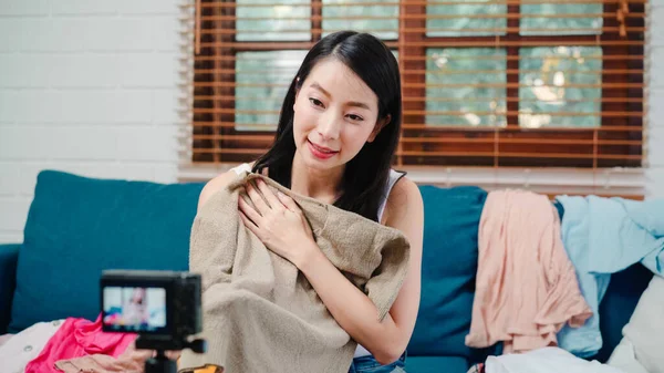 Asian stylist fashion influencer designer women using camera streaming and live to sell clothes, product, and accessories business, e-commerce broadcast online on sofa in living room at home.