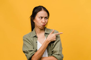 Genç Asyalı kadın, boş alanda inanılmaz bir şey gösteriyor, olumsuz ifadeli, heyecanlı çığlıklar atan, ağlayan, sinirli, sarı arka planda izole edilmiş günlük kıyafetlerle. Yüz ifadesi kavramı.