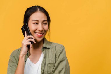 Genç Asyalı bayan telefonda olumlu bir ifadeyle konuşuyor, geniş bir şekilde gülümsüyor, gündelik giysiler içinde mutluluğu hissediyor ve sarı arka planda tek başına duruyor. Mutlu, mutlu, mutlu kadın başarıya sevinir..