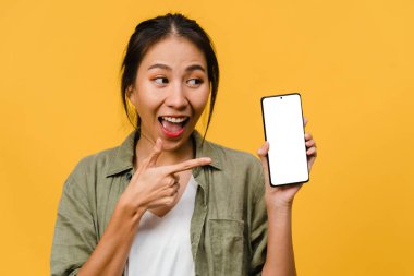 Genç Asyalı bayan boş akıllı telefon ekranını pozitif bir ifadeyle gösteriyor, geniş bir şekilde gülümsüyor, gündelik kıyafetler giyip sarı arka planda mutluluğu hissediyor. Kadın eli beyaz ekranlı cep telefonu..