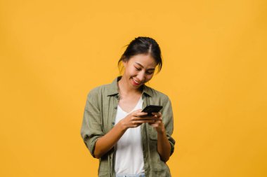 Genç Asyalı bayan telefonu olumlu bir ifadeyle kullanıyor, geniş bir şekilde gülümsüyor, gündelik giysiler içinde mutluluğu hissediyor ve sarı arka planda tek başına duruyor. Mutlu, mutlu, mutlu kadın başarıya sevinir..