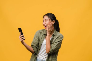 Genç Asyalı bayan telefonu olumlu bir ifadeyle kullanıyor, geniş bir şekilde gülümsüyor, gündelik giysiler içinde mutluluğu hissediyor ve sarı arka planda tek başına duruyor. Mutlu, mutlu, mutlu kadın başarıya sevinir..