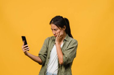 Asyalı genç bir bayanın telefonu olumlu bir ifadeyle kullandığını hayal etmek, günlük giysiler içinde mutluluğu hissetmek ve sarı arka planda izole bir şekilde durmak. Mutlu, mutlu, mutlu kadın başarıya sevinir..