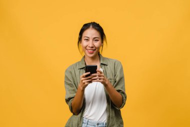 Asyalı genç bayanın cep telefonunu olumlu bir ifadeyle kullanmasına, geniş bir şekilde gülümsemesine, günlük kıyafetler giymesine ve sarı arka plandaki kameraya bakmasına şaşırdım. Mutlu, mutlu, mutlu kadın başarıya sevinir..