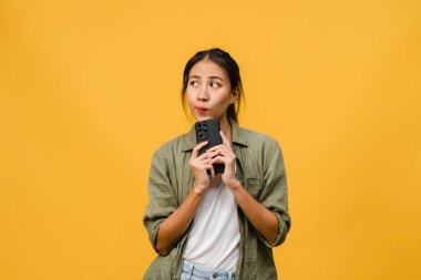 Asyalı genç bir bayanın telefonu olumlu bir ifadeyle kullandığını hayal etmek, günlük giysiler içinde mutluluğu hissetmek ve sarı arka planda izole bir şekilde durmak. Mutlu, mutlu, mutlu kadın başarıya sevinir..