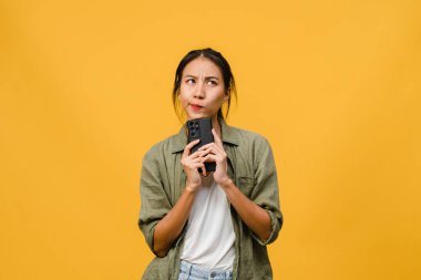 Asyalı genç bir bayanın telefonu olumlu bir ifadeyle kullandığını hayal etmek, günlük giysiler içinde mutluluğu hissetmek ve sarı arka planda izole bir şekilde durmak. Mutlu, mutlu, mutlu kadın başarıya sevinir..
